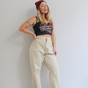 May include: A woman wearing a black crop top with the text "Harley Davidson" and a red beanie. She is wearing beige high-waisted jeans and black heeled sandals.