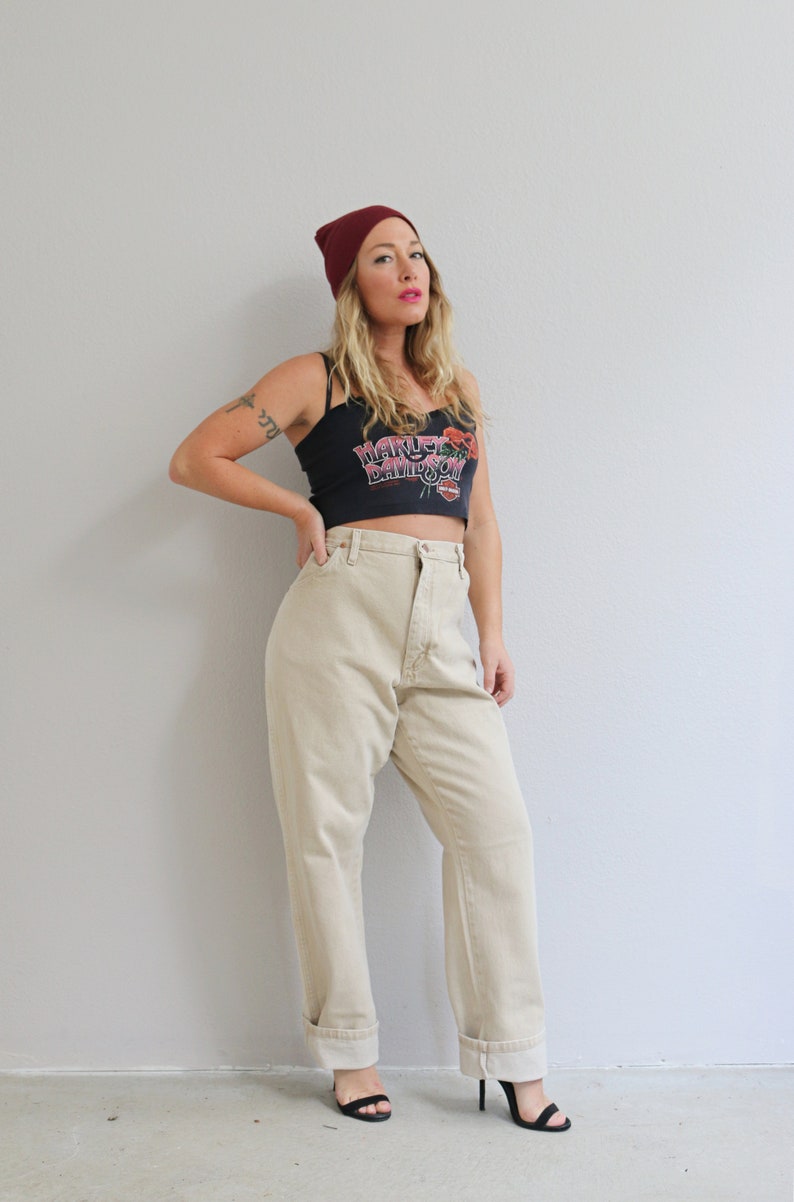 May include: A woman wearing a black crop top with the text "Harley Davidson" and a red beanie. She is wearing beige high-waisted jeans and black heeled sandals.