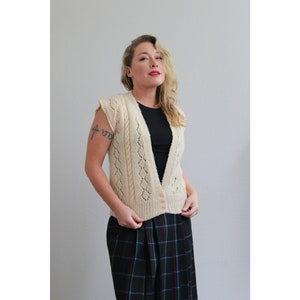 May include: Cream-colored cable knit vest with a V-neck and small buttons. The sleeveless sweater features a diamond pattern and is worn over a black top. The person is wearing a plaid skirt.