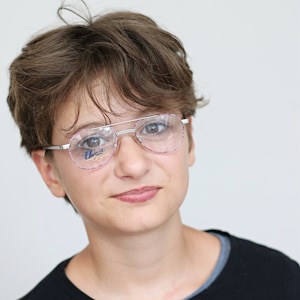 May include: A young person with short brown hair wearing silver framed glasses with pink accents. The person is wearing a black t-shirt.