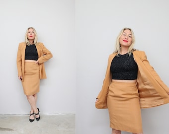 May include: A woman wearing a camel-colored linen suit with a black crop top. The suit includes a blazer and a skirt. She is wearing black heels.