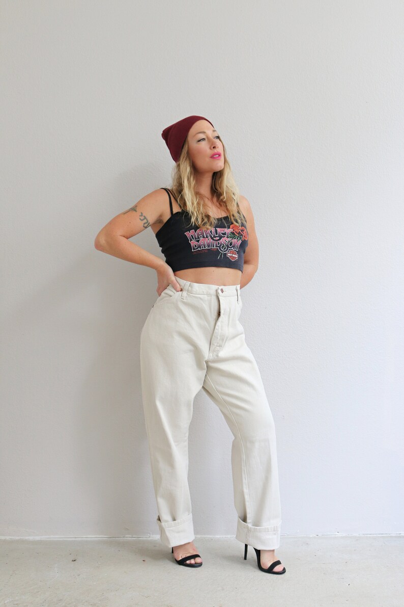 Pu&ograve; includere: Una donna indossa un top corto nero con la scritta "Harley Davidson" stampata, un berretto bordeaux, jeans bianchi a vita alta e sandali neri con tacco.