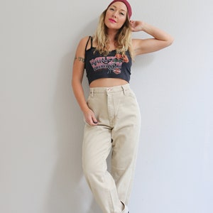 May include: A woman wearing a black crop top with a red "Harley Davidson" logo, beige high-waisted pants, and a burgundy beanie. She is standing in front of a white wall.