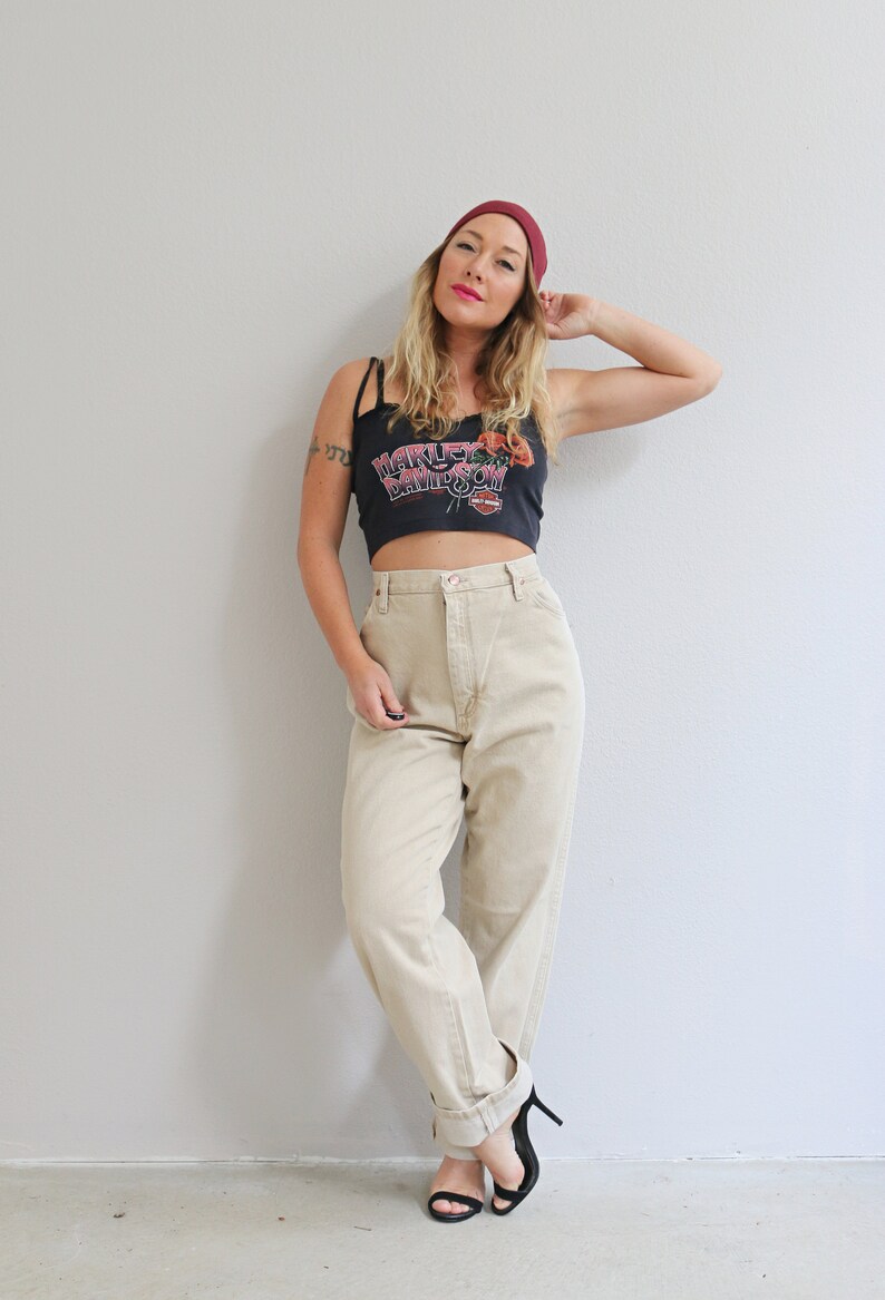 May include: A woman wearing a black crop top with a red "Harley Davidson" logo, beige high-waisted pants, and a burgundy beanie. She is standing in front of a white wall.
