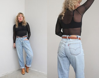 May include: A woman wearing a black long-sleeve mesh top, light blue high-waisted jeans, and a brown belt. The jeans have a classic fit. The woman is also wearing brown boots.