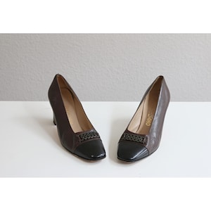 May include: A pair of brown leather pumps with black cap toes. The shoes have a decorative metal detail across the vamp and a medium-height heel. The shoes are displayed on a white surface.