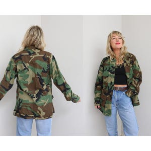 Puede incluir: Chaqueta con estampado de camuflaje en tonos verdes, marrones y negros. La chaqueta tiene cierre de botones en la parte delantera, mangas largas y múltiples bolsillos. La chaqueta se usa con vaqueros azules.