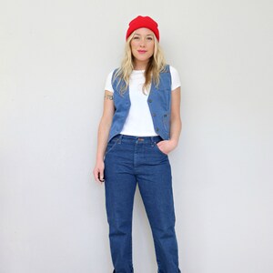 May include: A woman wearing a white t-shirt, a blue denim vest, blue jeans, and red shoes. She is also wearing a red beanie.