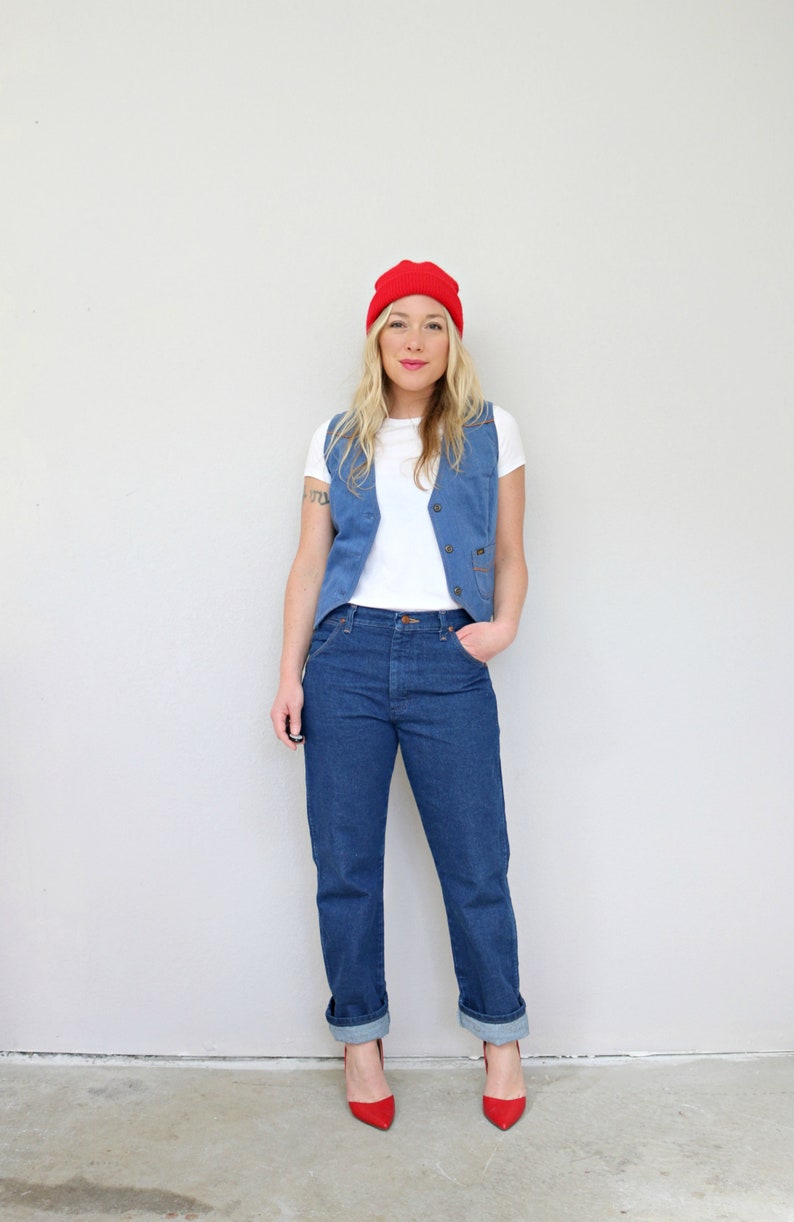 May include: A woman wearing a white t-shirt, a blue denim vest, blue jeans, and red shoes. She is also wearing a red beanie.