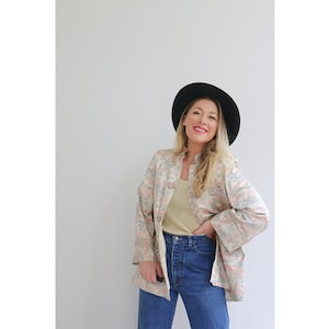 Puede incluir: Una mujer con una chaqueta de estampado floral, un top de tirantes marrón claro, pantalones vaqueros azules y un sombrero fedora negro.