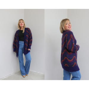 Puede incluir: Un cárdigan largo y extragrande con un patrón de chevrón. El cárdigan tiene una base de color púrpura oscuro con detalles en rojo, naranja y verde. La modelo lleva el cárdigan con una camiseta negra y vaqueros azules.