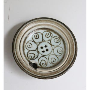 May include: Ceramic lid with a light blue interior featuring a brown spiral design and three small holes. The lid has a brown and beige rim and a small notch on the side.