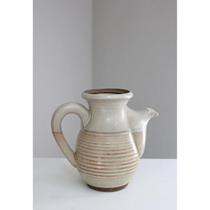 May include: A ceramic pitcher with a handle and spout. The pitcher is primarily off-white with horizontal brown stripes around the middle. The handle and spout have a brown accent. The pitcher sits on a white surface.