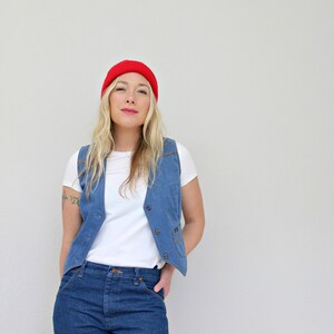 May include: A woman wearing a red beanie, a white t-shirt, a blue denim vest, and blue jeans.