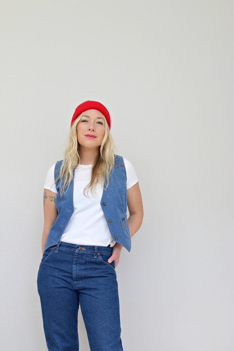May include: A woman wearing a red beanie, a white t-shirt, a blue denim vest, and blue jeans.