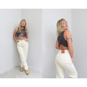 May include: A dark gray sleeveless crop top with a Rolling Stones graphic and cream-colored high-waisted jeans. The jeans have a classic five-pocket design and a red Levi's tag on the back pocket. Tan boots complete the look.