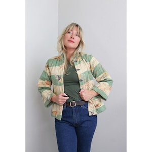 May include: A quilted jacket featuring horizontal stripes in alternating shades of sage green and cream. The fabric is decorated with a repeating pattern of small, stylized floral motifs in orange and brown. It has long sleeves and toggle closures.