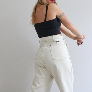 Pu&ograve; includere: Una donna indossa un top corto nero e jeans a vita alta color crema. I jeans hanno una tasca posteriore con una piccola toppa in pelle che dice "Wrangler".