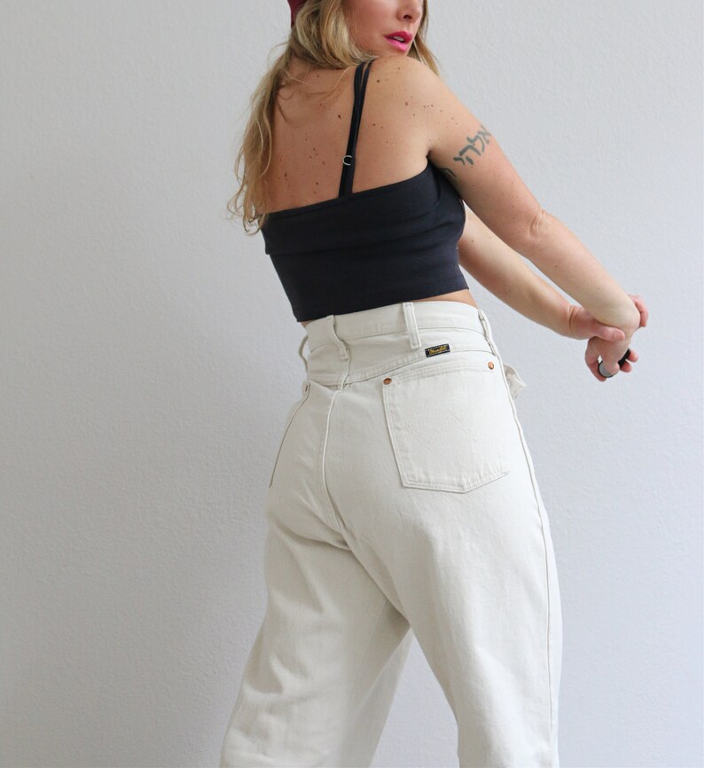 Pu&ograve; includere: Una donna indossa un top corto nero e jeans a vita alta color crema. I jeans hanno una tasca posteriore con una piccola toppa in pelle che dice "Wrangler".