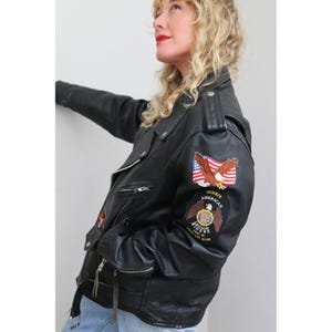May include: Black leather motorcycle jacket with multiple zippers, a belt, and patches. The patches include an American flag and an eagle emblem. The jacket has a classic biker style with a notched collar and shoulder epaulettes.