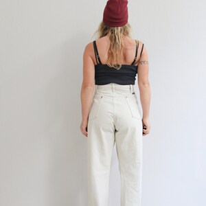 Pu&ograve; includere: Una persona indossa un paio di jeans bianchi a vita alta con orli arrotolati, una canotta nera e un berretto bordeaux.