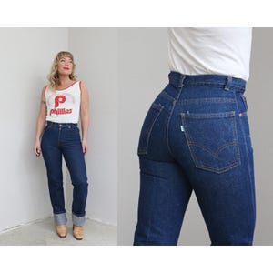 May include: Dark wash denim jeans with a high waist and straight leg. The jeans have a classic five-pocket design and a button and zipper closure. The jeans are cuffed at the ankles.
