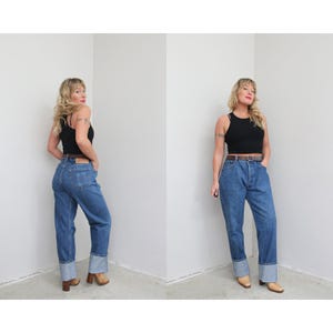 May include: Two images of a person wearing a black vest top, blue jeans with a brown belt, and brown shoes. The jeans have a cuffed hem. The person is standing in front of a white wall.