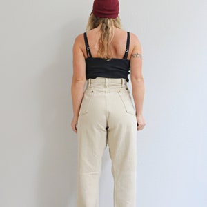 May include: A person wearing a burgundy beanie, a black tank top, and high-waisted, light beige jeans. The jeans have a cuffed hem.