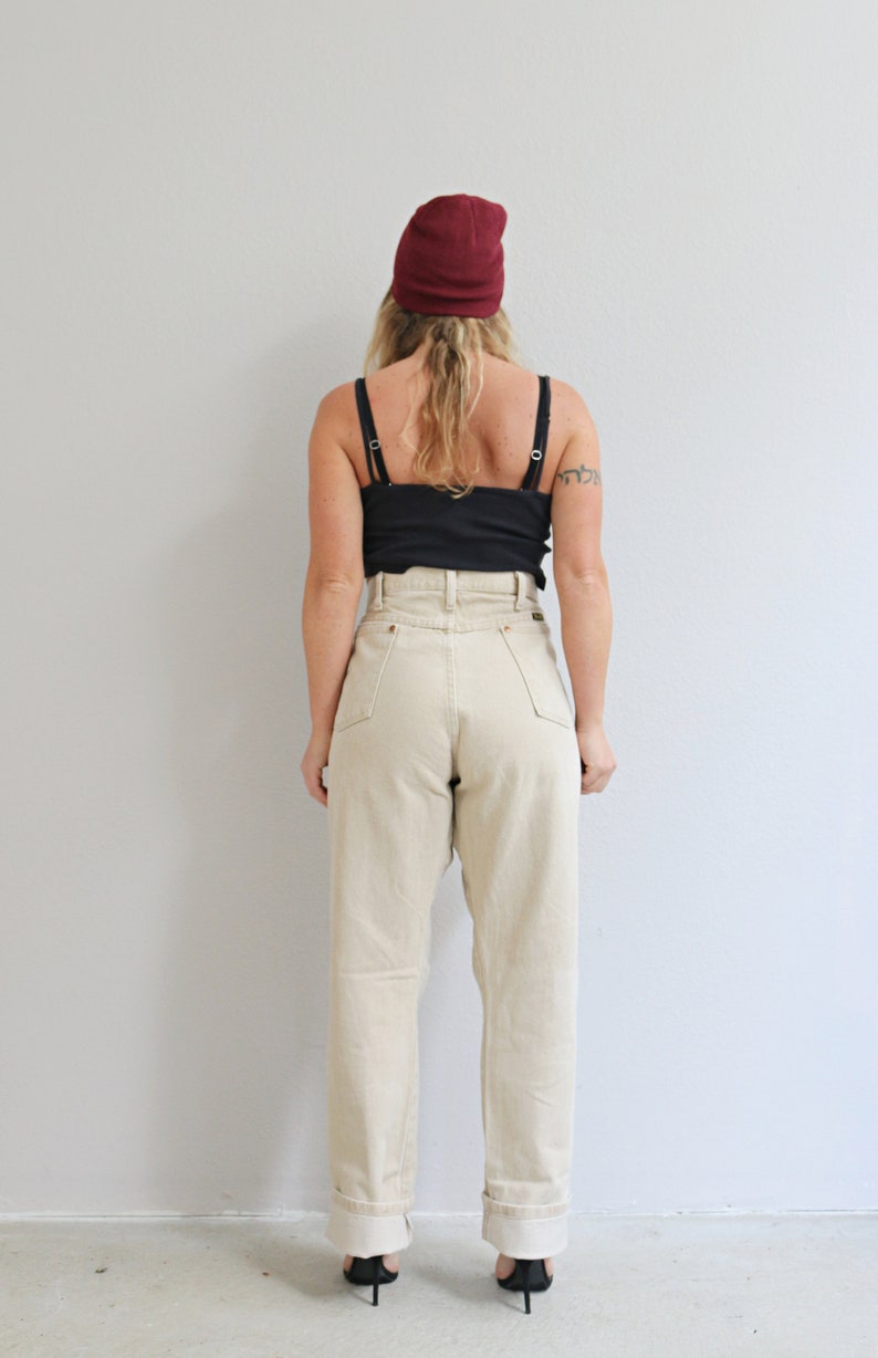 May include: A person wearing a burgundy beanie, a black tank top, and high-waisted, light beige jeans. The jeans have a cuffed hem.