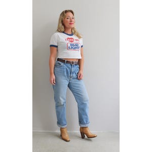 May include: A white crop top with a blue trim and the text "I love my Bud Light" in red and blue. The top is paired with light blue jeans, a brown belt, and tan heeled boots. The outfit is casual and vintage-inspired.