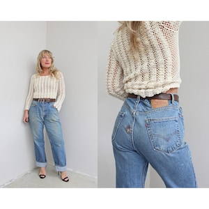 May include: A cream-colored, open-weave crochet sweater with long sleeves and a brown leather belt paired with light blue denim jeans. The jeans have a high waist and a straight leg fit.