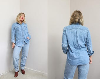 May include: A light blue denim button-down shirt with long sleeves, a pointed collar, and two chest pockets with buttoned flaps. The shirt features a classic western-style yoke across the shoulders and is paired with matching light blue denim jeans.