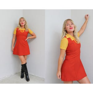 Puede incluir: Un vestido de estilo vintage con una parte superior amarilla con cuello y mangas cortas y un vestido rojo sin mangas. El vestido presenta un pequeño logotipo en el pecho. La modelo lleva botas negras hasta la rodilla.