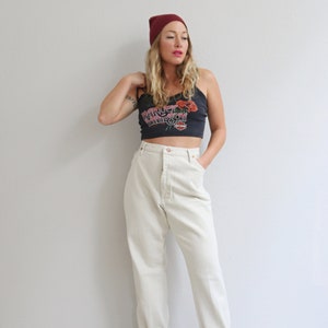 Pu&ograve; includere: Una donna indossa un berretto bordeaux, una canotta nera con un motivo floreale e la scritta "Harley Davidson" stampata, pantaloni beige chiaro a vita alta e sandali neri con tacco.