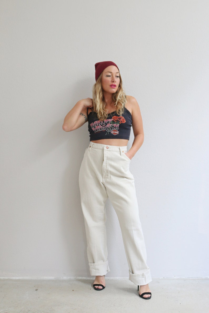 Pu&ograve; includere: Una donna indossa un berretto bordeaux, una canotta nera con un motivo floreale e la scritta "Harley Davidson" stampata, pantaloni beige chiaro a vita alta e sandali neri con tacco.