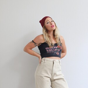 May include: A woman wearing a burgundy beanie, a black tank top with a Harley Davidson logo, and beige high-waisted jeans.