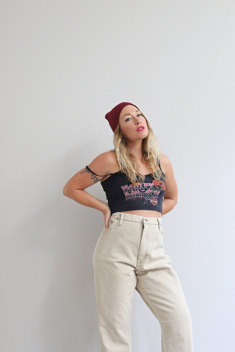 May include: A woman wearing a burgundy beanie, a black tank top with a Harley Davidson logo, and beige high-waisted jeans.