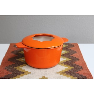 May include: Bright orange enameled cast iron fondue pot with a matching lid. The pot has a white interior and two side handles. It sits on a patterned, multi-colored textile.