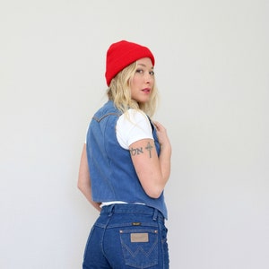 May include: A woman wearing a red beanie, a white t-shirt, a blue denim vest, and blue jeans. The jeans have a brown leather patch with the Wrangler logo on the back pocket.