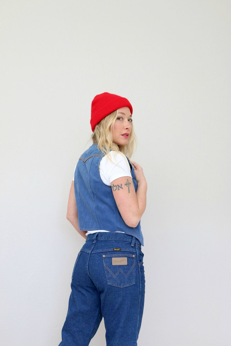 May include: A woman wearing a red beanie, a white t-shirt, a blue denim vest, and blue jeans. The jeans have a brown leather patch with the Wrangler logo on the back pocket.