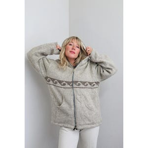May include: A light gray, long-sleeved, zip-up hooded jacket featuring a horizontal band across the chest with a repeating brown wave pattern. The jacket has a front zipper closure and two open pockets at the waist, crafted from a textured knit fabric.