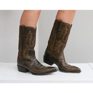 May include: A pair of dark brown leather cowboy boots featuring a pointed toe, a stacked heel, and decorative stitching patterns on the shaft. The boots have a pull-on design with side tabs and a distressed finish on the leather surface.