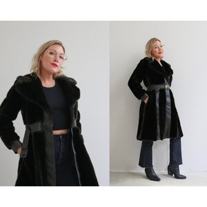 May include: A long black faux fur coat with a leather belt and a black leather panel down the front. The coat has a wide collar and is worn by a person with blonde hair.
