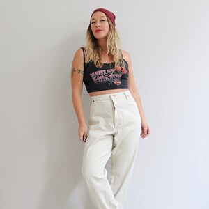 Pu&ograve; includere: Una donna indossa una canotta nera con il logo Harley Davidson e fiori rossi, jeans bianchi a vita alta e un berretto bordeaux.