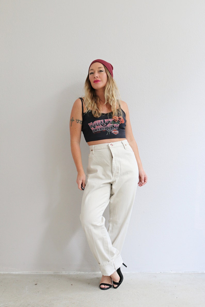 Pu&ograve; includere: Una donna indossa una canotta nera con il logo Harley Davidson e fiori rossi, jeans bianchi a vita alta e un berretto bordeaux.
