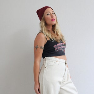 Pu&ograve; includere: Una donna indossa un berretto bordeaux, una canotta nera con la scritta "Harley Davidson" stampata e jeans bianchi. Ha un tatuaggio di una croce sul braccio.