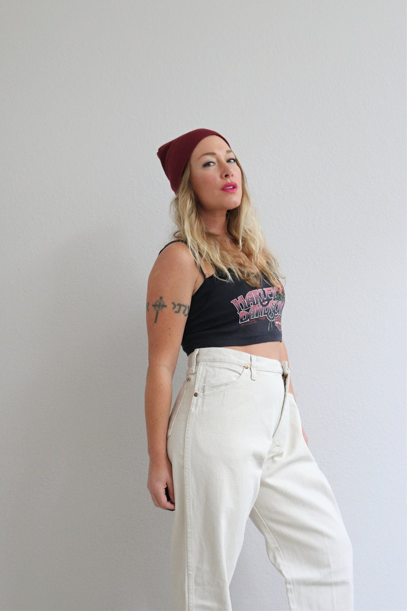 Pu&ograve; includere: Una donna indossa un berretto bordeaux, una canotta nera con la scritta "Harley Davidson" stampata e jeans bianchi. Ha un tatuaggio di una croce sul braccio.