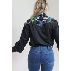 May include: A long-sleeved black button-down shirt featuring a western-style yoke with a multi-colored floral and checkered pattern in shades of purple, green, yellow, and white, paired with blue denim jeans with a visible Wrangler brand label.