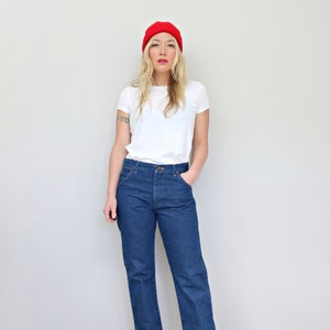 May include: A woman wearing a white t-shirt, blue jeans, and a red beanie. She is standing in front of a white wall and is looking at the camera.