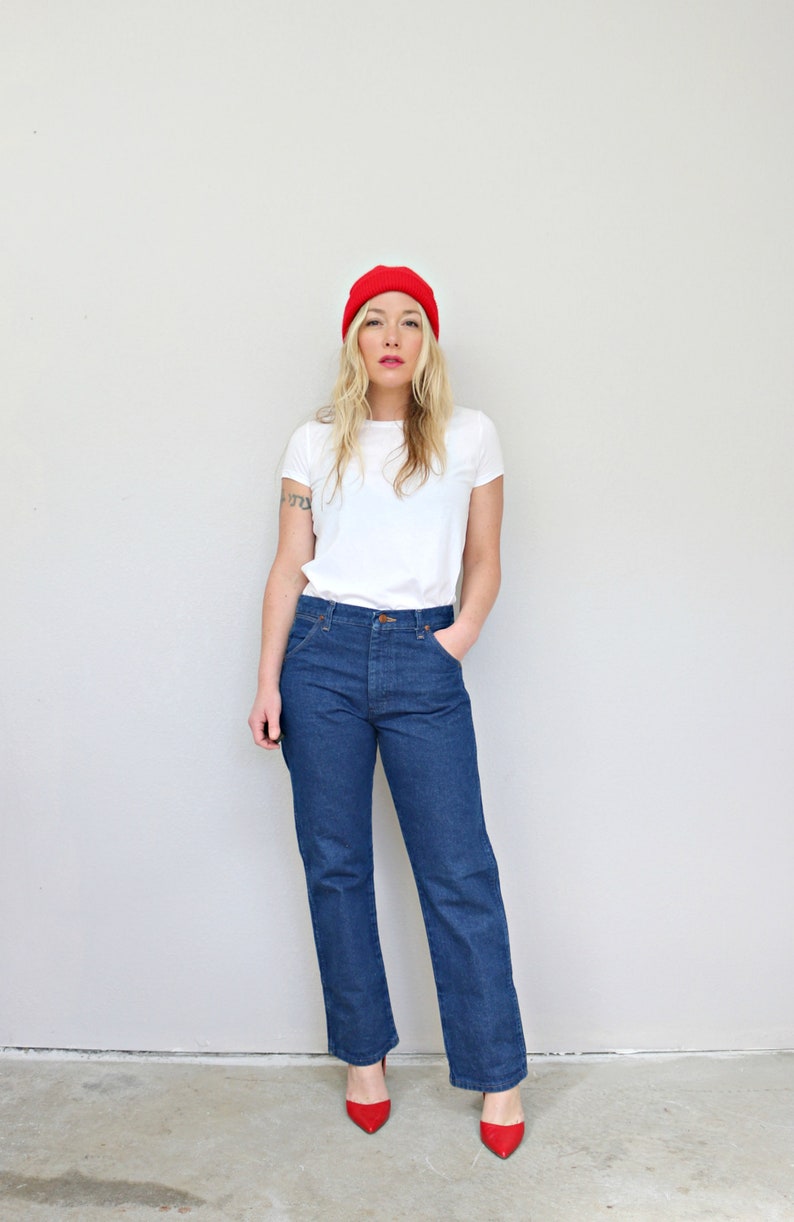 May include: A woman wearing a white t-shirt, blue jeans, and a red beanie. She is standing in front of a white wall and is looking at the camera.